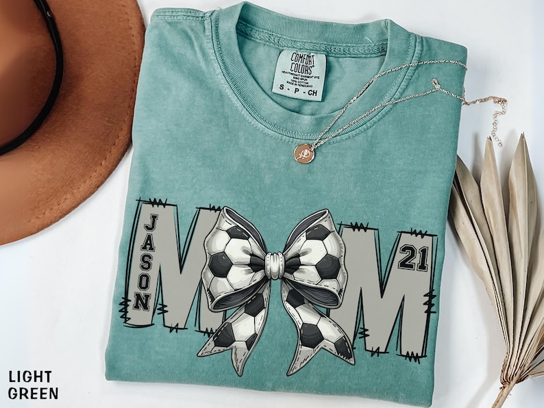 May include: Light green t-shirt with a large white and black bow with a soccer ball pattern. The bow is centered on the shirt with the letters "MOM" on either side. The left side of the shirt has the name "JASON" and the right side has the number "21".