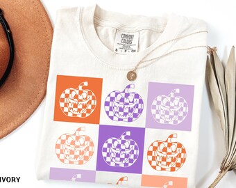 May include: Ivory-colored t-shirt featuring a grid pattern of jack-o'-lanterns with checkered orange and purple designs. The shirt has a Comfort Colors label and a gold necklace. The word "IVORY" is printed at the bottom.