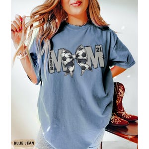 May include: A blue jean t-shirt with the word "MOM" in white letters with a soccer ball bow in the middle. The text "JASON 21" is on the shirt.