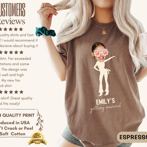 May include: A brown t-shirt with a white graphic of a woman wearing sunglasses and dancing. The text "EMILY'S getting married" is printed below the graphic. The shirt is being modeled by a person.