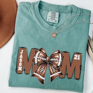 May include: Light green t-shirt with a football-themed design. The word "MOM" is displayed in brown letters, with a football bow in the center. The name "JASON" and the number "21" are also visible. A gold necklace is draped across the shirt.