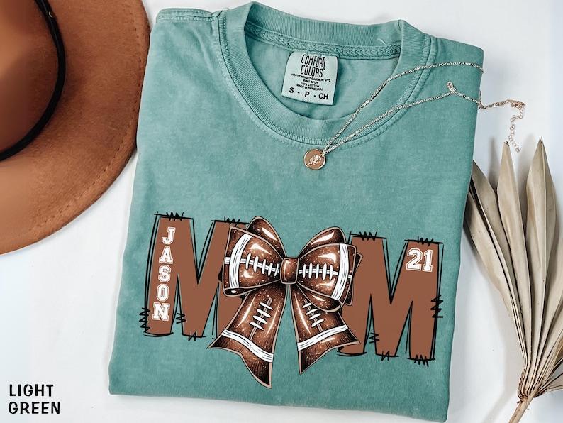 May include: Light green t-shirt with a football-themed design. The word "MOM" is displayed in brown letters, with a football bow in the center. The name "JASON" and the number "21" are also visible. A gold necklace is draped across the shirt.