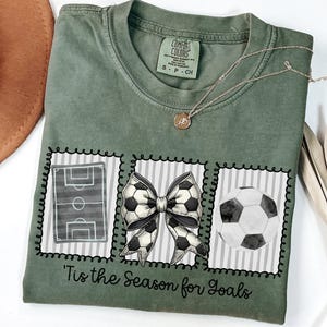 Puede incluir: Camiseta verde musgo con tem&aacute;tica de f&uacute;tbol. La camiseta presenta tres im&aacute;genes enmarcadas: un campo de f&uacute;tbol, un lazo de bal&oacute;n de f&uacute;tbol y un bal&oacute;n de f&uacute;tbol. El texto "'Tis the Season for Goals" est&aacute; impreso debajo.