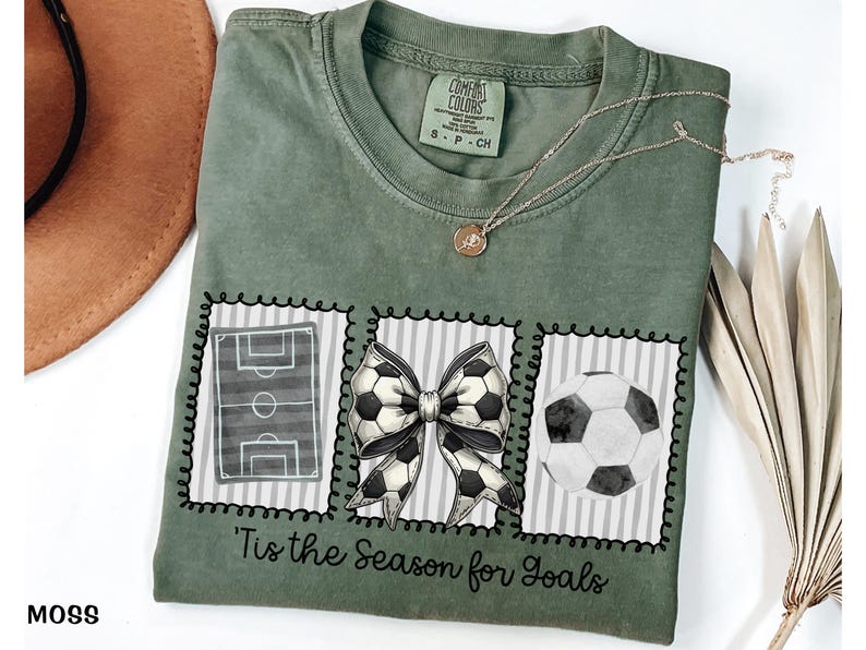 Puede incluir: Camiseta verde musgo con tem&aacute;tica de f&uacute;tbol. La camiseta presenta tres im&aacute;genes enmarcadas: un campo de f&uacute;tbol, un lazo de bal&oacute;n de f&uacute;tbol y un bal&oacute;n de f&uacute;tbol. El texto "'Tis the Season for Goals" est&aacute; impreso debajo.