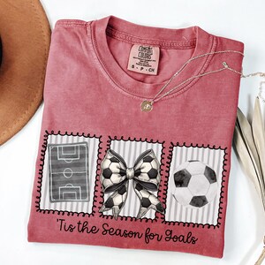 Puede incluir: Camiseta roja carmes&iacute; con tem&aacute;tica de f&uacute;tbol. La camiseta presenta un campo de f&uacute;tbol, un lazo de bal&oacute;n de f&uacute;tbol y un gr&aacute;fico de bal&oacute;n de f&uacute;tbol. El texto "'Tis the Season for Goals" est&aacute; impreso en la camiseta. Un collar plateado con un colgante est&aacute; en la camiseta.
