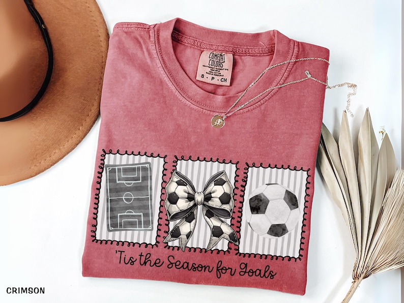 Puede incluir: Camiseta roja carmes&iacute; con tem&aacute;tica de f&uacute;tbol. La camiseta presenta un campo de f&uacute;tbol, un lazo de bal&oacute;n de f&uacute;tbol y un gr&aacute;fico de bal&oacute;n de f&uacute;tbol. El texto "'Tis the Season for Goals" est&aacute; impreso en la camiseta. Un collar plateado con un colgante est&aacute; en la camiseta.