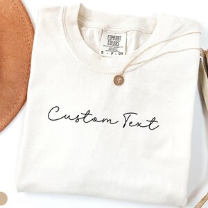 May include: Ivory colored t-shirt with the text "Custom Text" in black script font.