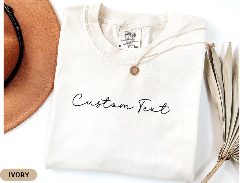 May include: Ivory colored t-shirt with the text "Custom Text" in black script font.