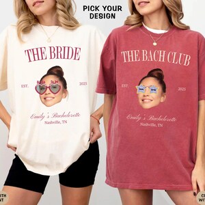 May include: Two white t-shirts with a red print. The text on the shirts reads "The Bride" and "The Bach Club". Both shirts feature a woman's face with sunglasses and the text "Emily's Bachelorette Nashville, TN".