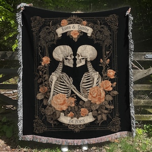 May include: Black throw blanket featuring a romantic skeleton couple design. The design includes two skeletons facing each other, surrounded by roses, with the names "Raven & Damien" and the year "Est 2028" on banners. The blanket has a fringed edge.