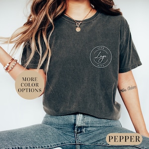 May include: A dark gray t-shirt with a white circle on the left chest that says "Your Logo Here". The shirt is being worn by a person with long brown hair and blue jeans. The text "Pepper" is visible on a brown tag.