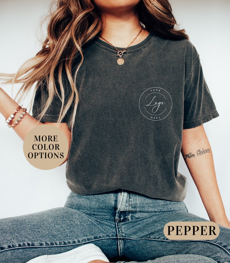 May include: A dark gray t-shirt with a white circle on the left chest that says "Your Logo Here". The shirt is being worn by a person with long brown hair and blue jeans. The text "Pepper" is visible on a brown tag.