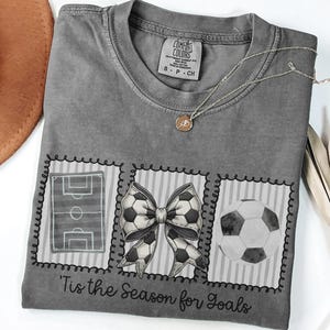 Puede incluir: Camiseta gris con tem&aacute;tica de f&uacute;tbol. La camiseta presenta tres dise&ntilde;os cuadrados: un campo de f&uacute;tbol, un lazo hecho con balones de f&uacute;tbol y un bal&oacute;n de f&uacute;tbol. El texto "'Tis the Season for Goals" est&aacute; impreso debajo.