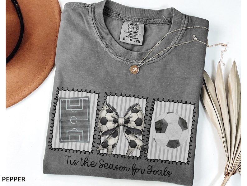 Puede incluir: Camiseta gris con tem&aacute;tica de f&uacute;tbol. La camiseta presenta tres dise&ntilde;os cuadrados: un campo de f&uacute;tbol, un lazo hecho con balones de f&uacute;tbol y un bal&oacute;n de f&uacute;tbol. El texto "'Tis the Season for Goals" est&aacute; impreso debajo.