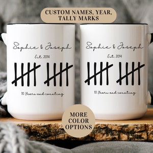 May include: Two white ceramic mugs with black handles and black rims. The mugs have black text that reads "Sophie & Joseph Est. 2014" and tally marks representing 10 years. The text below the tally marks reads "10 Years and counting".