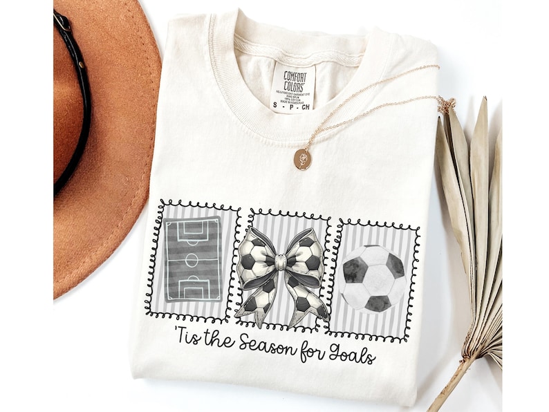 Puede incluir: Camiseta color crema con tem&aacute;tica de f&uacute;tbol. La camiseta presenta tres im&aacute;genes enmarcadas: un campo de f&uacute;tbol, un lazo con detalles de bal&oacute;n de f&uacute;tbol y un bal&oacute;n de f&uacute;tbol. El texto "'Tis the Season for Goals" est&aacute; debajo.