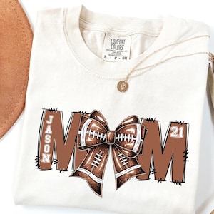 May include: Ivory-colored t-shirt with a football-themed design. The word "MOM" is displayed in brown block letters, with a football bow in the center. The name "JASON" and the number "21" are also featured.