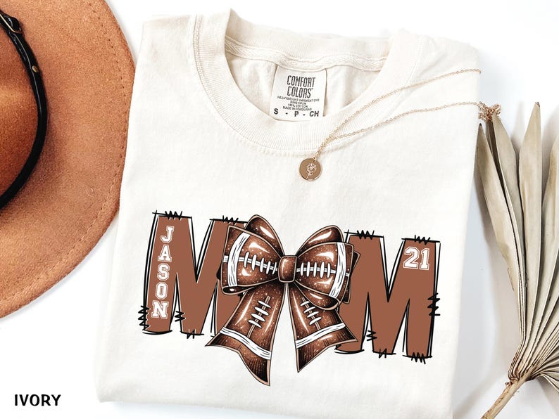 May include: Ivory-colored t-shirt with a football-themed design. The word "MOM" is displayed in brown block letters, with a football bow in the center. The name "JASON" and the number "21" are also featured.