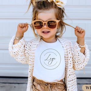 May include: A white t-shirt with a black circle and the word "Logo" written in cursive inside the circle. The shirt is being worn by a young child with blonde hair and brown sunglasses. The child is wearing a white cardigan and brown pants.