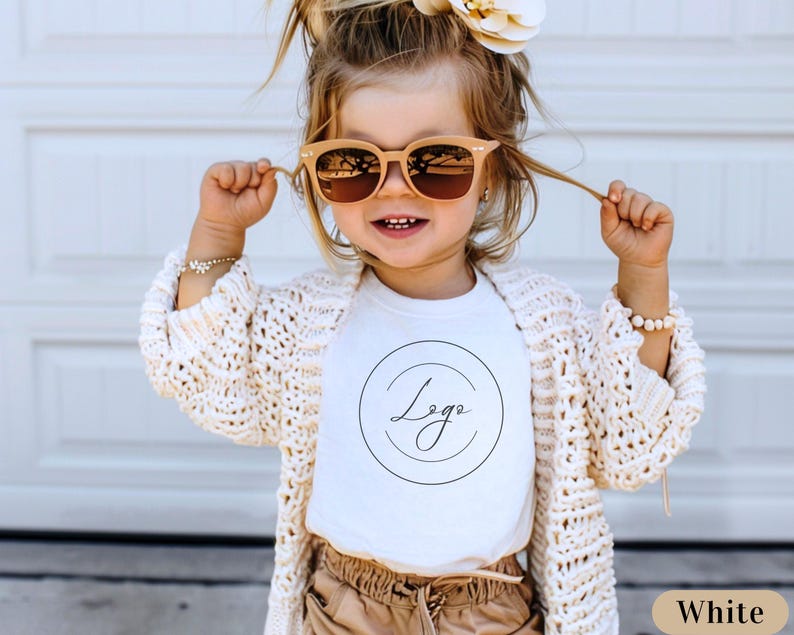 May include: A white t-shirt with a black circle and the word "Logo" written in cursive inside the circle. The shirt is being worn by a young child with blonde hair and brown sunglasses. The child is wearing a white cardigan and brown pants.