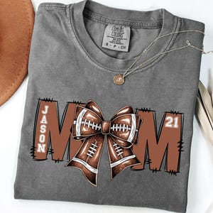May include: Gray t-shirt with a football-themed design. The design includes the word "MOM" with a football bow in the center, the name "JASON" on the left, and the number "21" on the right. A gold necklace is also visible.
