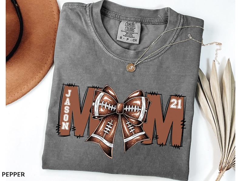 May include: Gray t-shirt with a football-themed design. The design includes the word "MOM" with a football bow in the center, the name "JASON" on the left, and the number "21" on the right. A gold necklace is also visible.