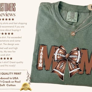 May include: Moss green t-shirt with a football-themed design. The word "MOM" is displayed in brown block letters, with a football bow in the center. The name "JASON" and the number "21" are also visible. A silver necklace is draped on the shirt.