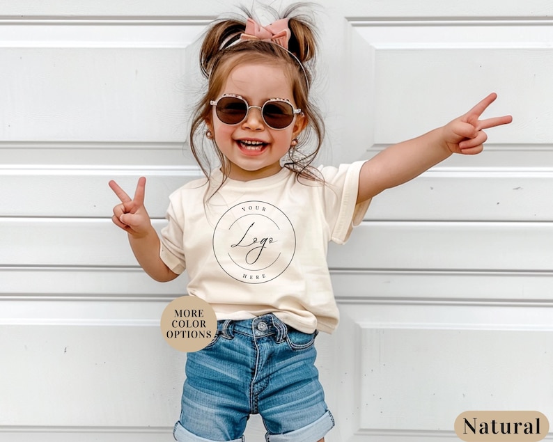 May include: A young child wearing a light beige t-shirt with a circle graphic that says "Your Logo Here" in black script. The child is wearing blue denim shorts and has a pink bow in their hair. The child is smiling and making peace signs with both hands. The background is a white wall.