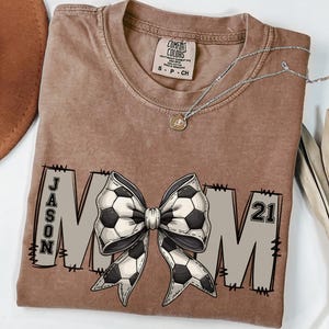 May include: A brown t-shirt with a white and black soccer ball bow and the text "JASON MOM 21" printed on it.