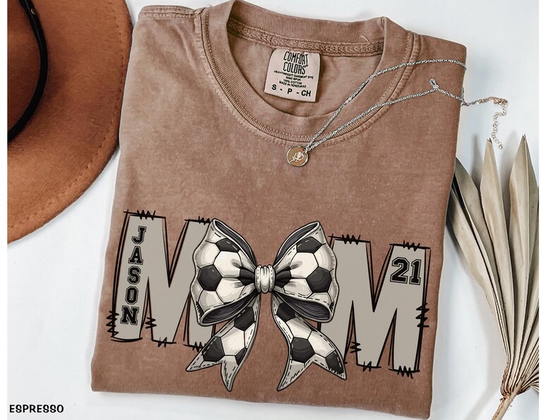 May include: A brown t-shirt with a white and black soccer ball bow and the text "JASON MOM 21" printed on it.