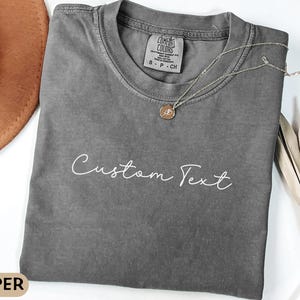May include: A gray,  comfy-looking t-shirt with the text "Custom Text" printed in white on the front. The shirt has a relaxed fit and a crew neck.