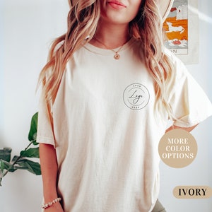 May include: A woman is wearing a light beige t-shirt with a black circle logo that says "Your Logo Here". The t-shirt is available in other colors. The text "More Color Options" and "Ivory" are also visible in the image.