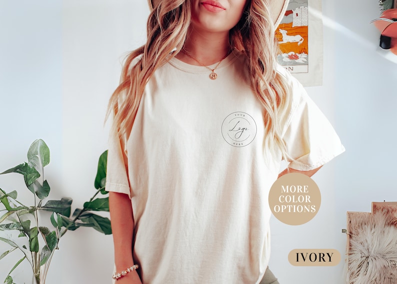 May include: A woman is wearing a light beige t-shirt with a black circle logo that says "Your Logo Here". The t-shirt is available in other colors. The text "More Color Options" and "Ivory" are also visible in the image.