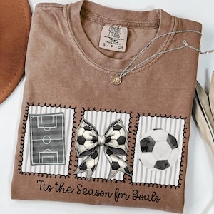 Puede incluir: Camiseta color espresso con tem&aacute;tica de f&uacute;tbol. La camiseta presenta tres im&aacute;genes enmarcadas: un campo de f&uacute;tbol, un lazo de bal&oacute;n de f&uacute;tbol y un bal&oacute;n de f&uacute;tbol. El texto "'Tis the Season for Goals" est&aacute; impreso debajo.