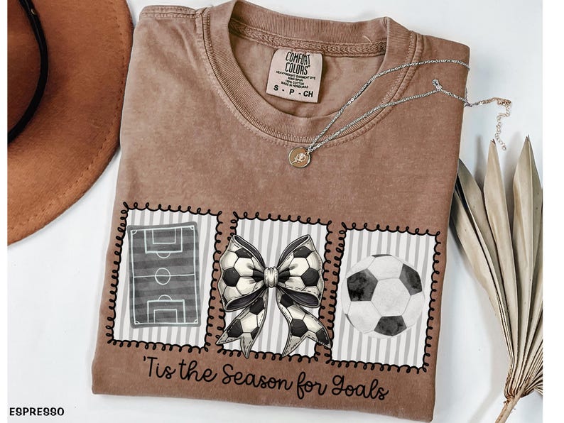 Puede incluir: Camiseta color espresso con tem&aacute;tica de f&uacute;tbol. La camiseta presenta tres im&aacute;genes enmarcadas: un campo de f&uacute;tbol, un lazo de bal&oacute;n de f&uacute;tbol y un bal&oacute;n de f&uacute;tbol. El texto "'Tis the Season for Goals" est&aacute; impreso debajo.