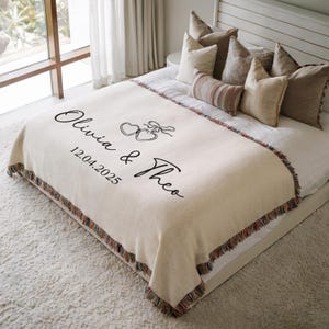 May include: Cream-colored throw blanket with fringed edges, personalized with the names "Olivia & Theo" and the date "12.04.2025" in black script. The blanket features a heart design and is displayed on a bed with pillows.