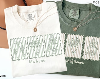 May include: Two t-shirts, one ivory and one moss green, with matching graphic designs. Each shirt features a series of three postage stamp-style images: champagne flutes, a bouquet, and a champagne bottle. The ivory shirt says "the bride", and the green shirt says "maid of honor".