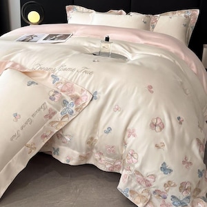 Pode incluir: Conjunto de roupa de cama creme com borboletas e flores bordadas em tons pastel. O conjunto inclui uma capa de edredão, fronhas e um lençol de cetim rosa. As palavras "Dreams Come True" são bordadas.