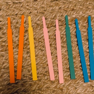 May include: Eight thin, flat strips of paper in various colors, including orange, yellow, pink, and green. The strips are arranged in a row on a woven surface.