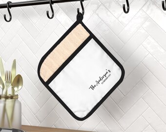 May include: White pot holder with black trim and a beige fabric insert. The pot holder has the text "The Anderson's Established 2021" printed on it.