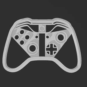May include: A light gray controller-shaped cookie cutter. The cutter features a detailed design of a game controller, including buttons, directional pad, and joysticks. The design is set against a dark gray background.