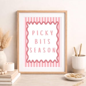 Könnte beinhalten: Ein gerahmter Druck mit dem Text "PICKY BITS SEASON" in Rosa. Der Hintergrund hat rosa und weiße vertikale Streifen. Der Rahmen ist hellbraun. Der Druck befindet sich auf einer weißen Oberfläche mit einer Vase, einer Kerze und anderen dekorativen Gegenständen.