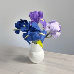 K&ouml;nnte beinhalten: Ein Strau&szlig; aus Filz-Iris in Blaut&ouml;nen und Lila, arrangiert in einer wei&szlig;en Keramikvase mit Punkten.