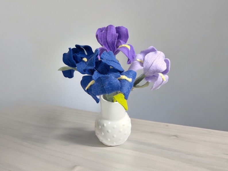 K&ouml;nnte beinhalten: Ein Strau&szlig; aus Filz-Iris in Blaut&ouml;nen und Lila, arrangiert in einer wei&szlig;en Keramikvase mit Punkten.