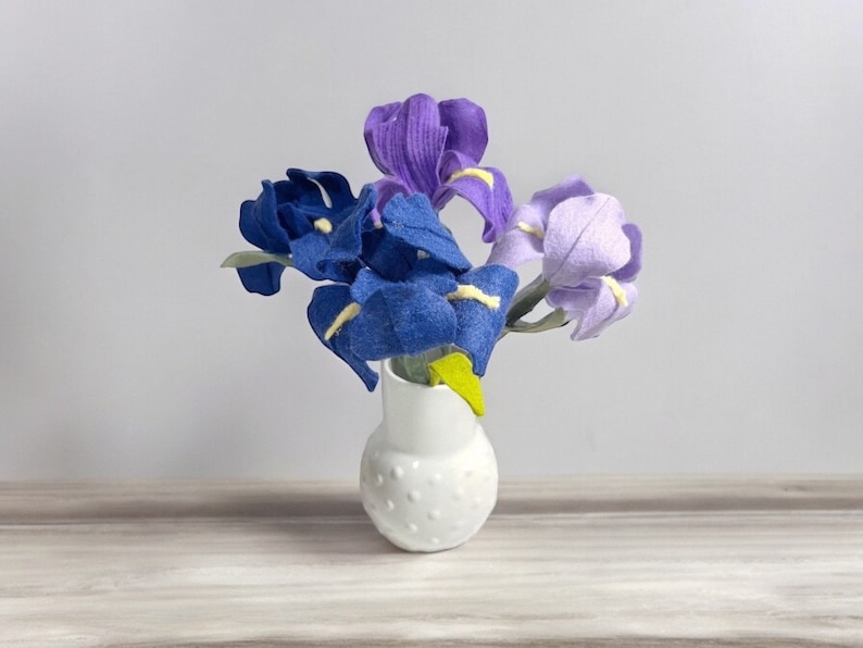 K&ouml;nnte beinhalten: Ein Strau&szlig; aus violetten, blauen und lavendelfarbenen Filz-Iris in einer wei&szlig;en Keramikvase mit Punkten.