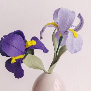 May include: Two felt iris flowers in a white vase. One flower is purple and the other is light purple. The flowers are on a light wood surface.