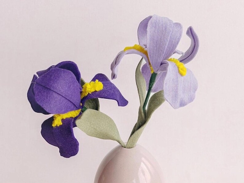 May include: Two felt iris flowers in a white vase. One flower is purple and the other is light purple. The flowers are on a light wood surface.