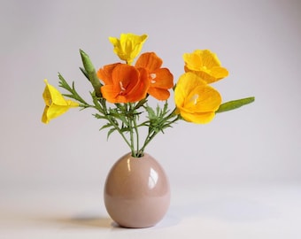 Puede incluir: Un ramo de flores de papel de color amarillo brillante y naranja con tallos y hojas verdes, dispuesto en un jarrón de cerámica de color rosa claro con forma de huevo. Las flores tienen pétalos delicados y arrugados con centros blancos.