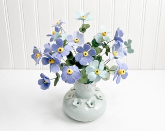 May include: A bouquet of light blue and periwinkle felt flowers with yellow centers and green felt leaves, arranged in a pale blue ceramic vase featuring a rounded base and small molded flower accents around the neck of the vessel.