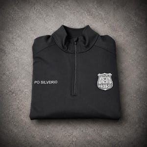 May include: Black quarter-zip pullover with the name "PO SILVERIO" embroidered on the left chest. A New York City Police Department badge with the number "23150" is on the right chest. The shirt is folded on a gray surface.